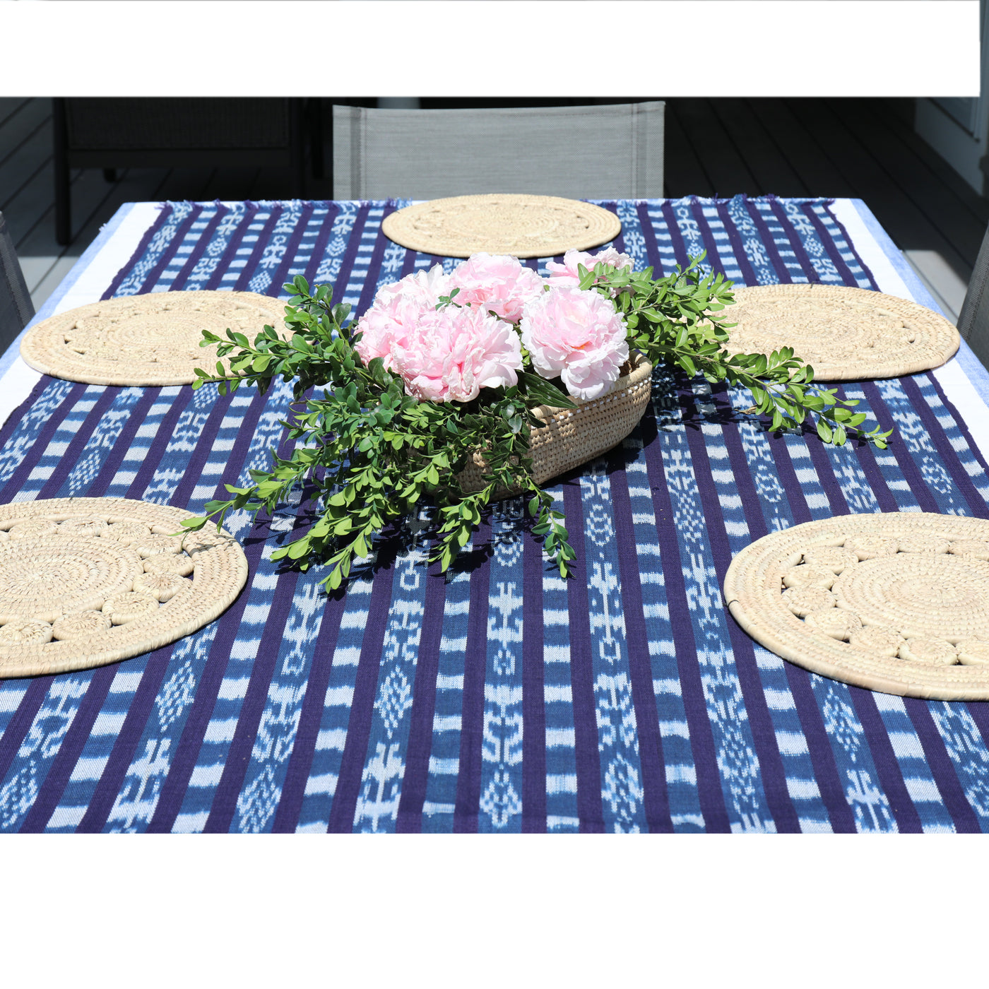 Table setting with a blue patterned tablecloth, woven placemats, and a floral centerpiece.