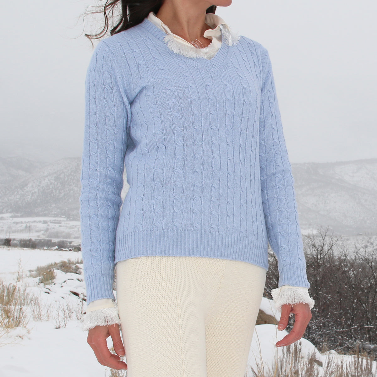 A woman wearing a light blue cable-knit sweater with white faux-fur trim at the collar and cuffs, paired with cream knit pants, standing in a snowy outdoor landscape.