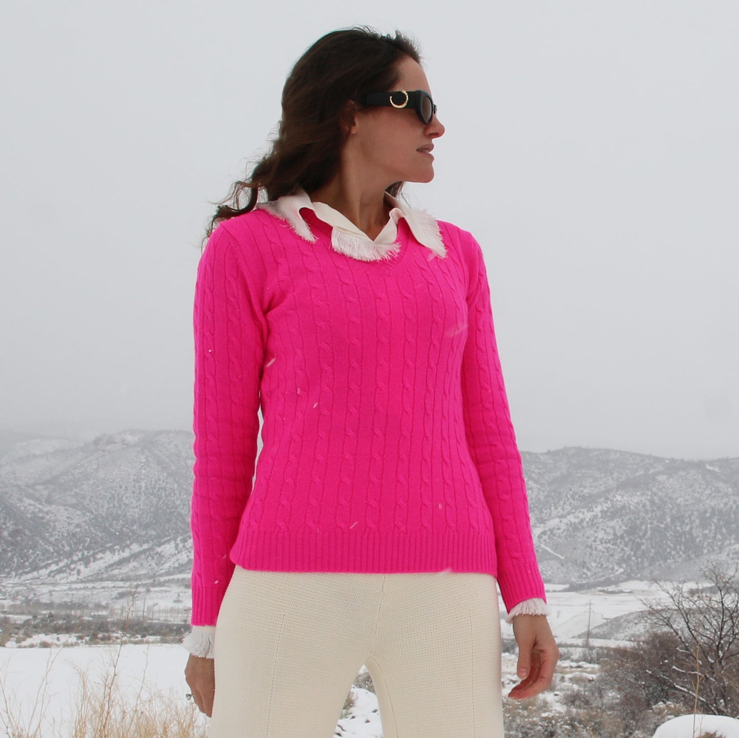 A woman wearing a bright pink cable-knit sweater with a white ruffled collar, paired with cream pants, standing outdoors in a snowy landscape.