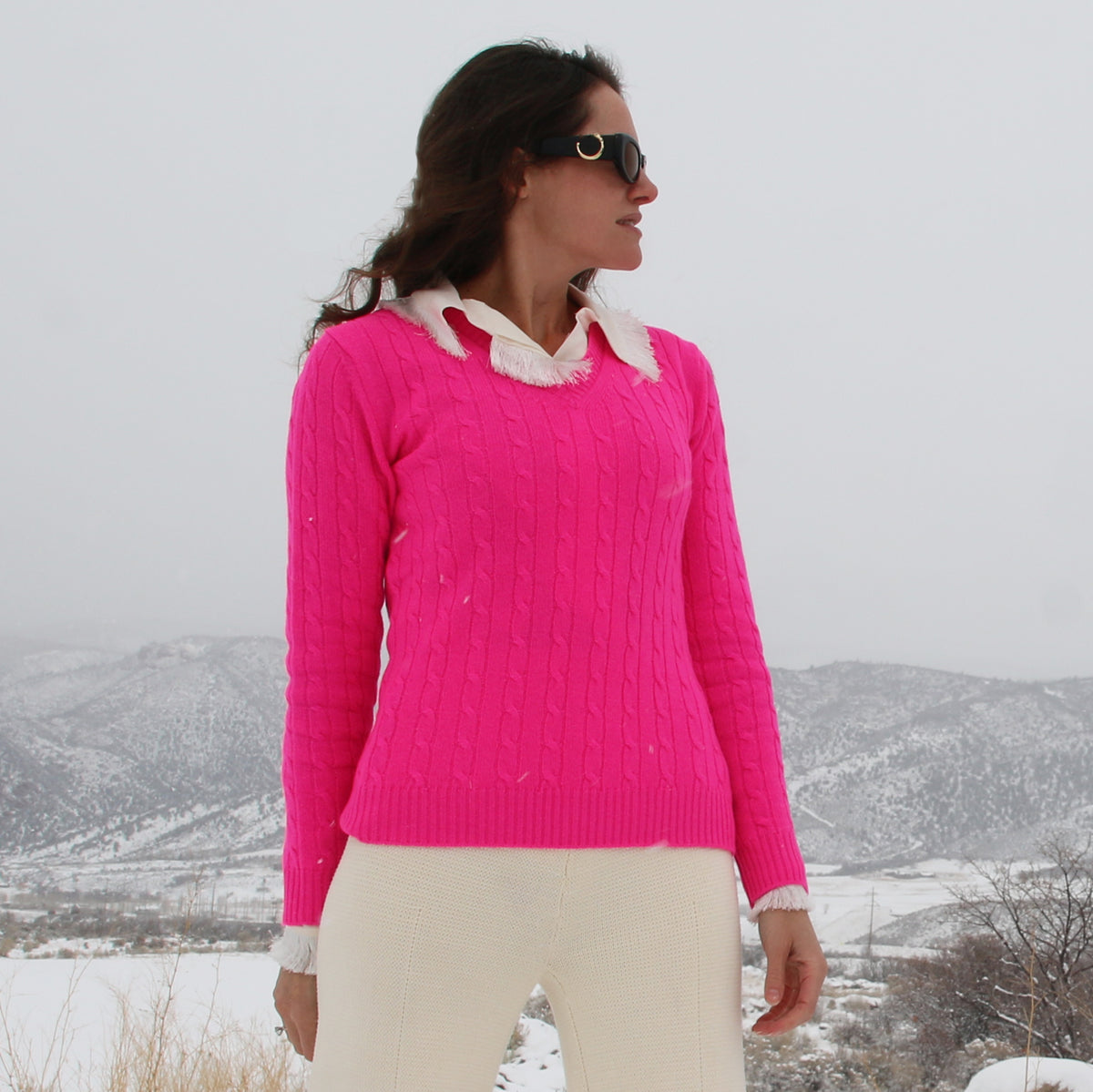 A woman wearing a bright pink cable-knit sweater with a white ruffled collar, paired with cream pants, standing outdoors in a snowy landscape.