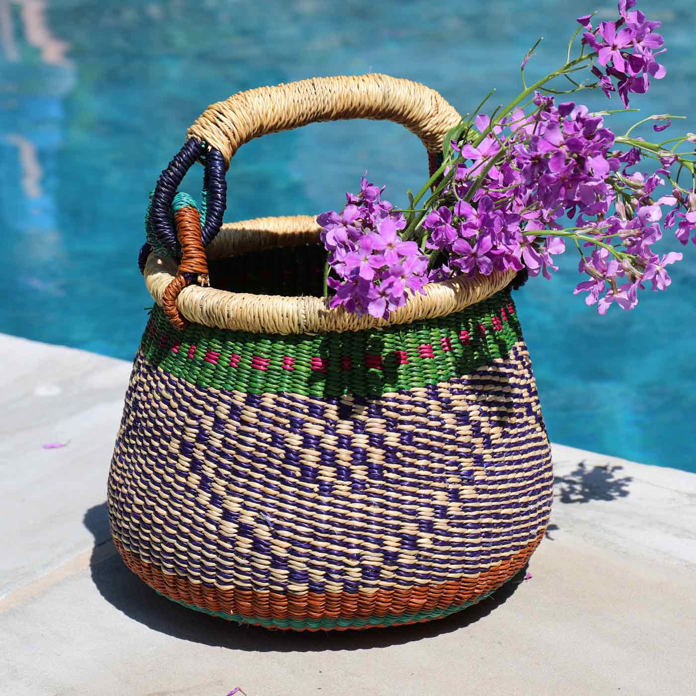 Woven basket with a handle filled with purple flowers by a pool.