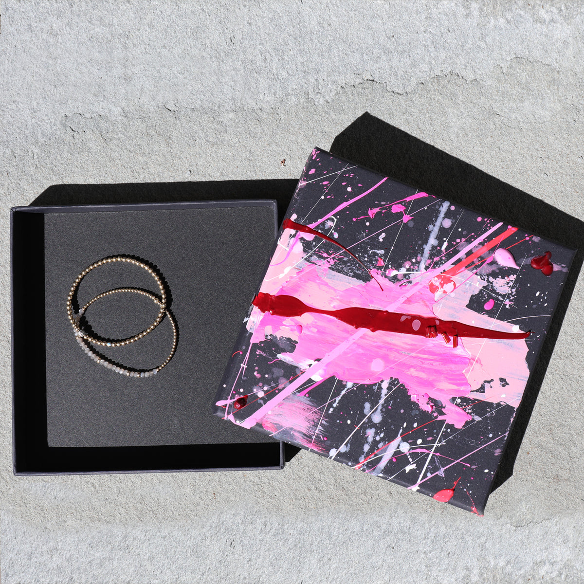 A pair of beaded rings in a textured black gift box next to a black lid decorated with a vibrant pink, red, and white paint splatter design, set on a light gray concrete surface.