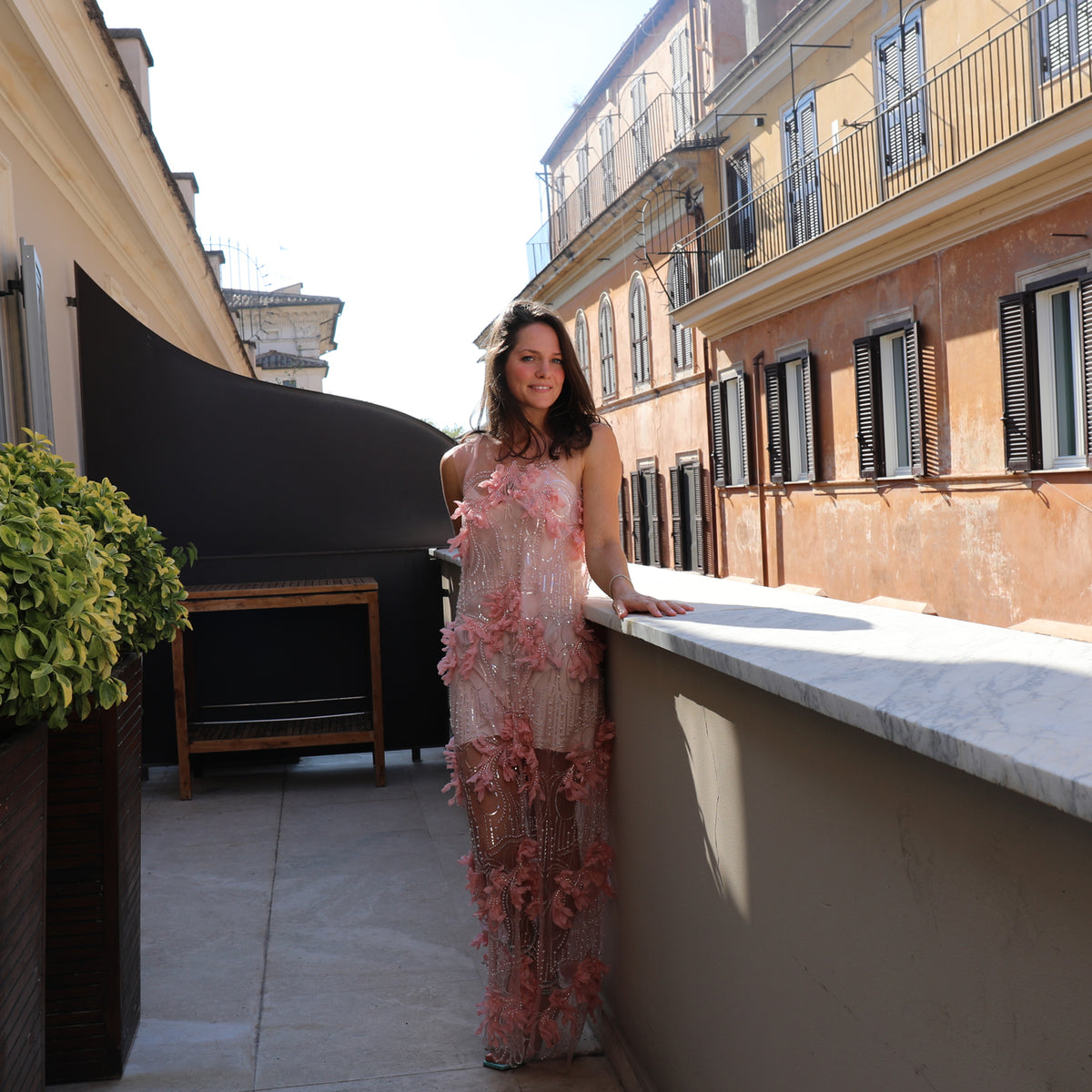 A woman wearing a pink floral lace dress stands on a sunlit balcony with a stone wall and Mediterranean-style buildings in the background.