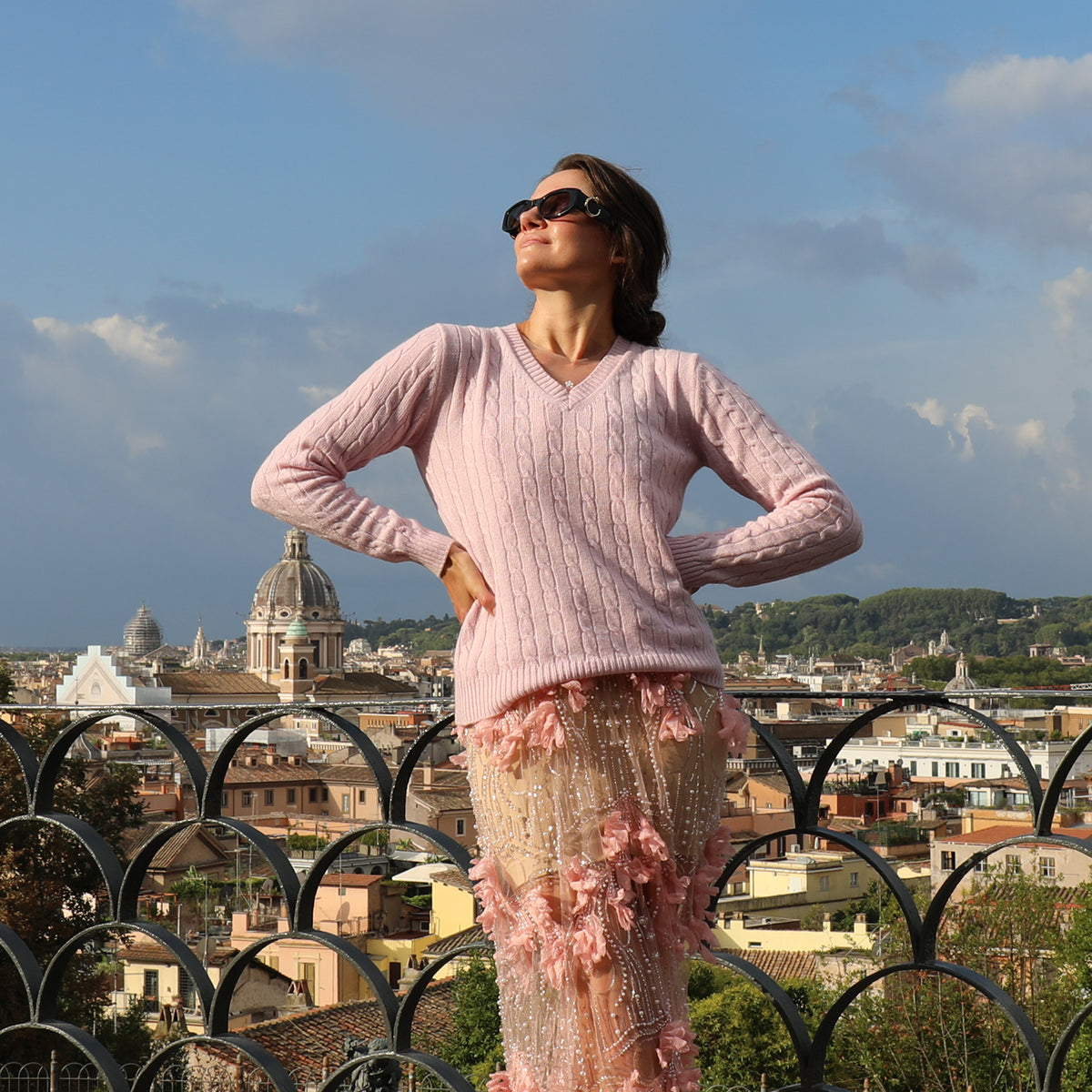 A woman wearing a light pink cable-knit sweater and a sheer pink embellished skirt stands outdoors on a balcony overlooking a cityscape with domes and hills under a blue sky.