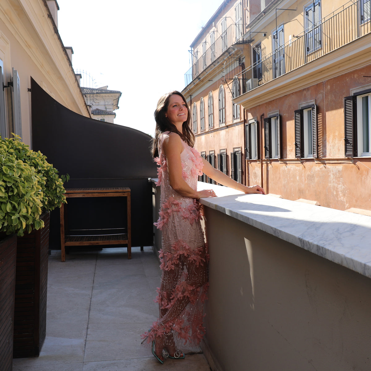 A woman wearing a pink floral lace maxi dress leans against a white marble balcony railing on a sunlit terrace with classic European buildings in the background.
