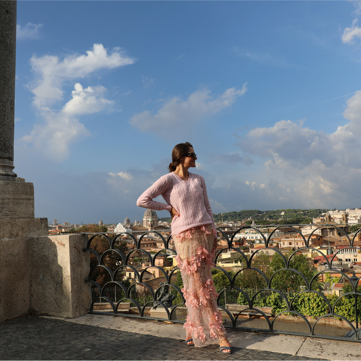 A woman stands on an outdoor balcony overlooking a cityscape under a blue sky, wearing a light pink knit sweater and a matching pink floral tulle skirt.