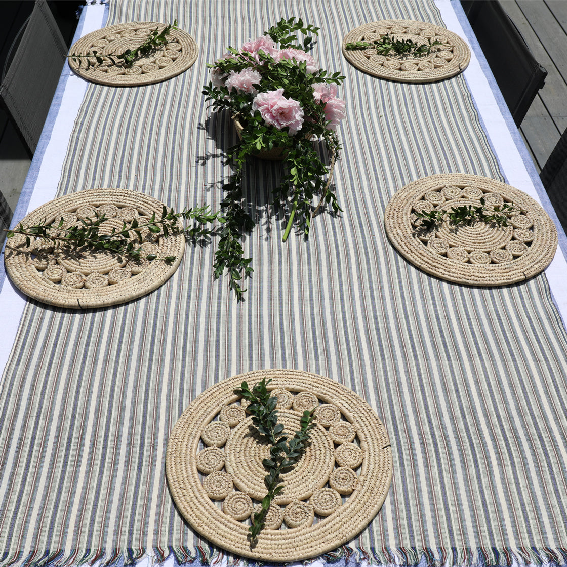 Set of round woven placemats on a striped tablecloth with a floral centerpiece.
